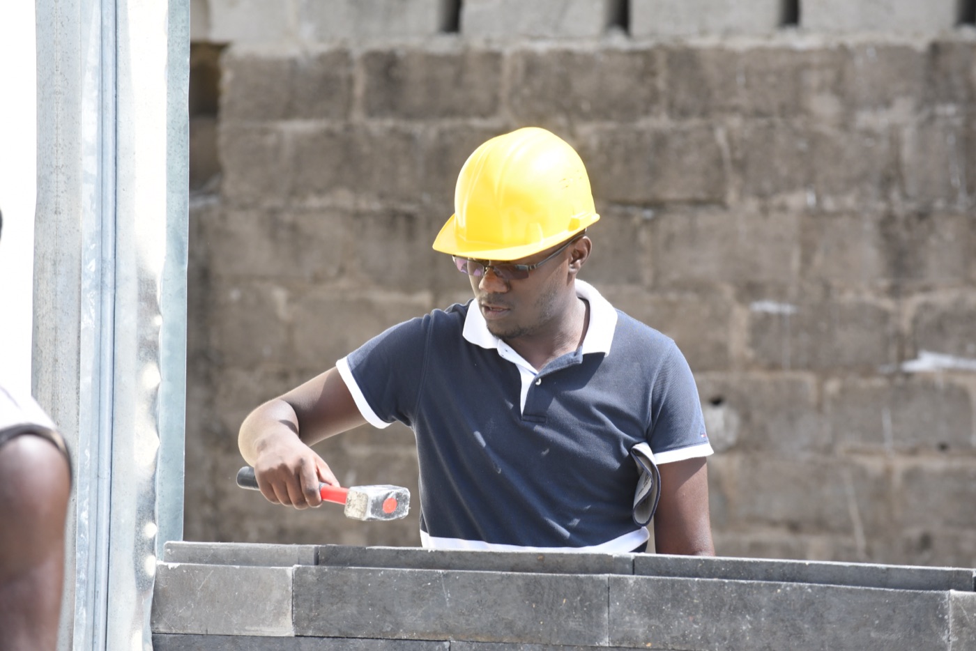A person wearing a yellow hard hat and safety glasses uses a rubber mallet to fit a recycled-plastic building block into place during construction of the first recycled-plastic classroom in Cote d'Ivoire.