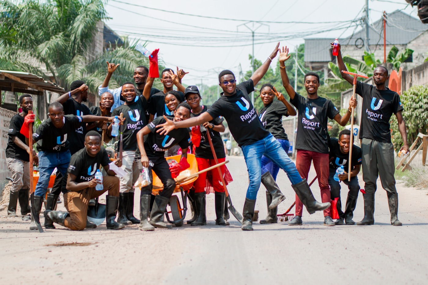 A group of U-Report DRC volunteers in matching black "U-Report par UNICEF" T-shirts, posing and cheering together on a street during a community clean-up.